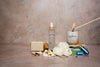 Collection of bath and body products including a candle, soap, room spray and inhaler on a textured surface.